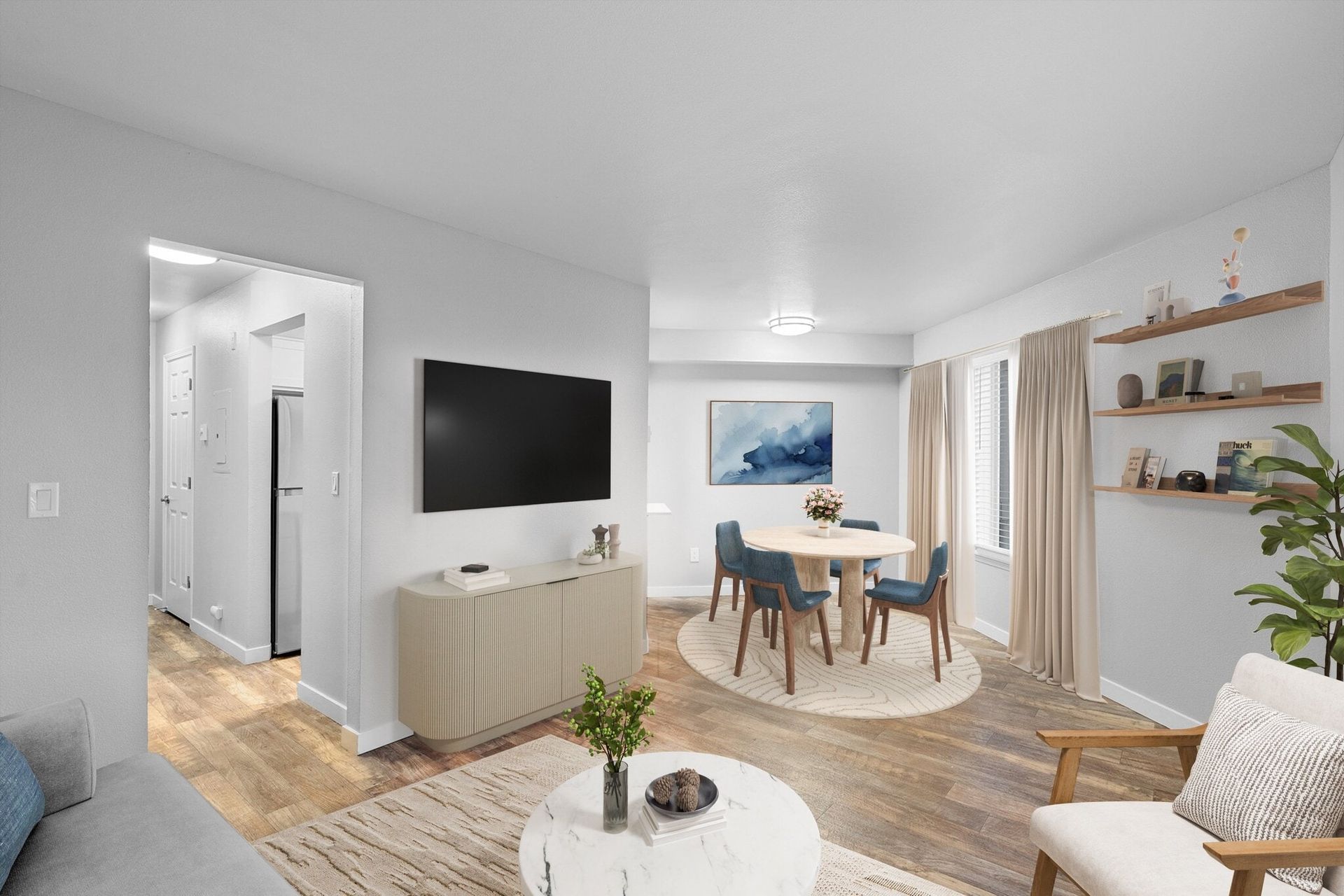 Living room with TV, dining area, and entrance to a hallway. Light walls, wood floors, and neutral furniture.