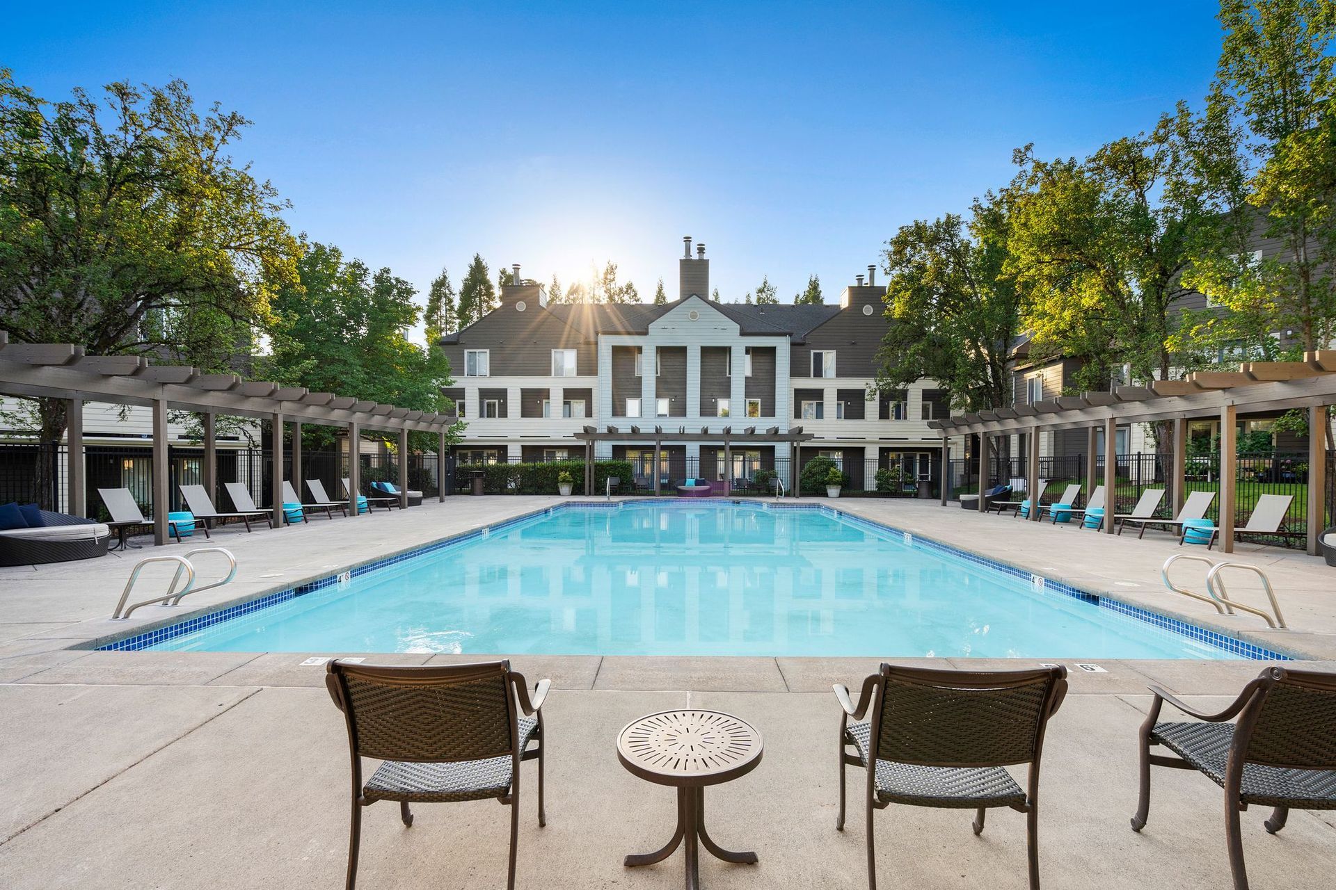 Swimming pool in front of a large, two-story building. Lounge chairs surround the pool with overhead pergolas. Blue sky.