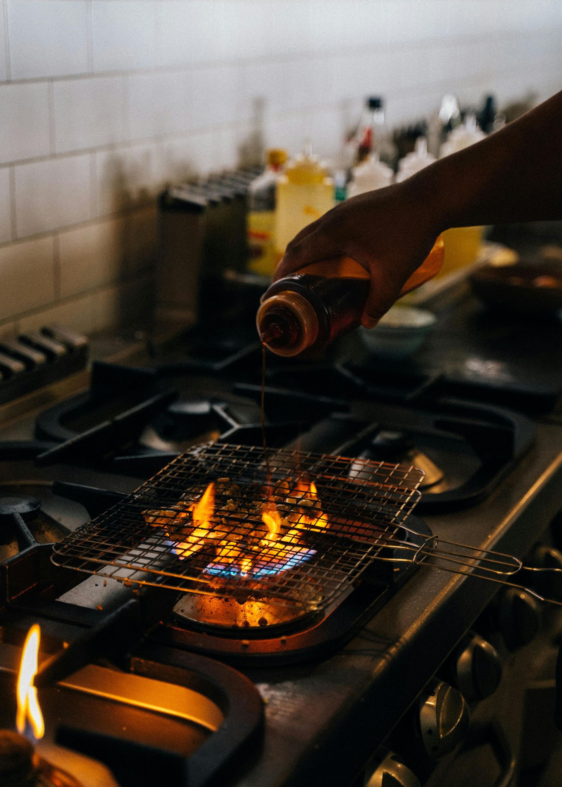 A Hand Pouring from A Bottle onto Food Grilling Over Flames on A Stovetop — Kuranda Gas & Supplies In Koah, QLD