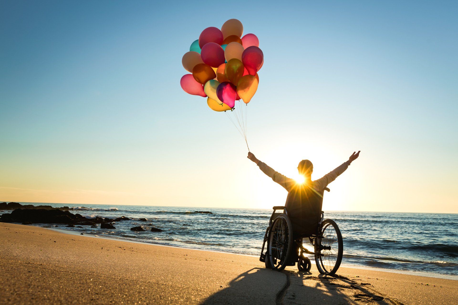 A person in a wheelchair is holding balloons on the beach.