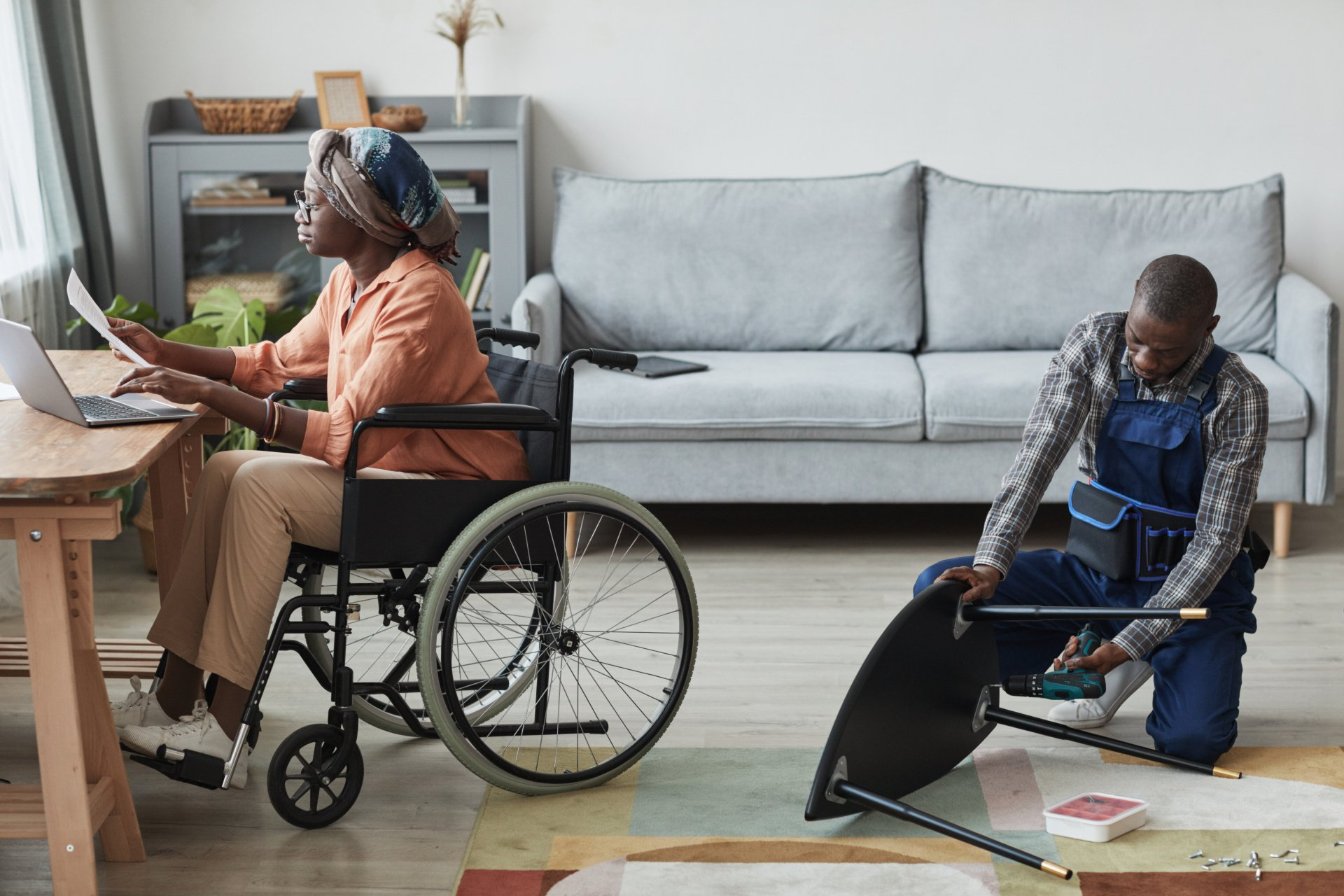 A woman in a wheelchair is sitting at a table while a man is kneeling down to fix a chair.