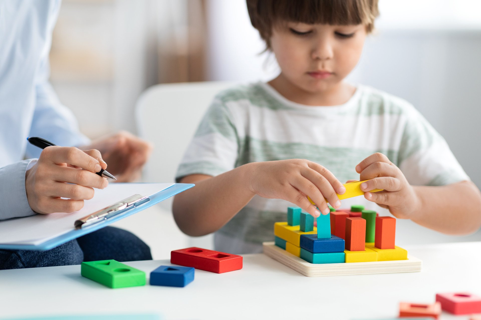 A young boy is playing with wooden blocks while a woman writes on a clipboard.