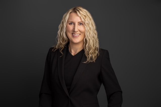 Woman with blonde curly hair smiles in a black suit. Dark background.