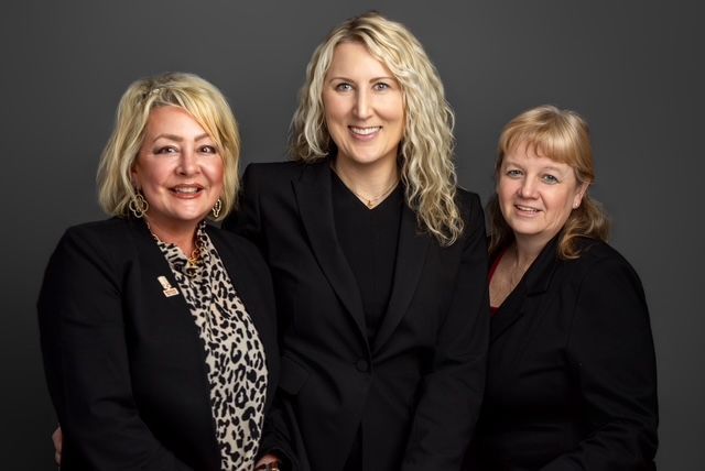 Three women in black blazers smile, posing against a dark gray backdrop.