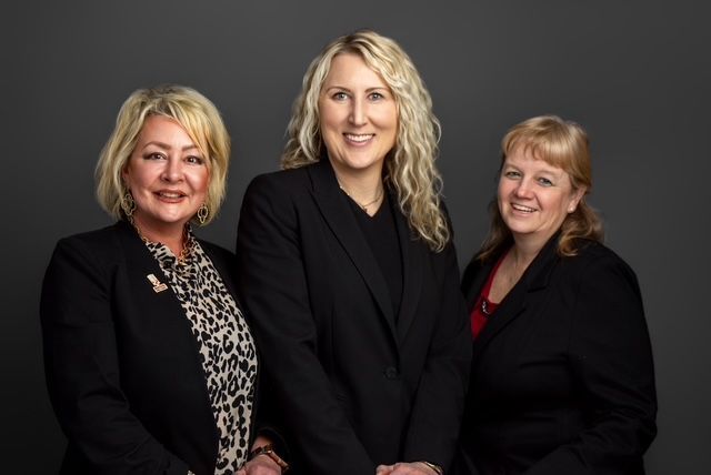 Three women in business attire, posing. Dark blazers, smiling, against a gray backdrop.