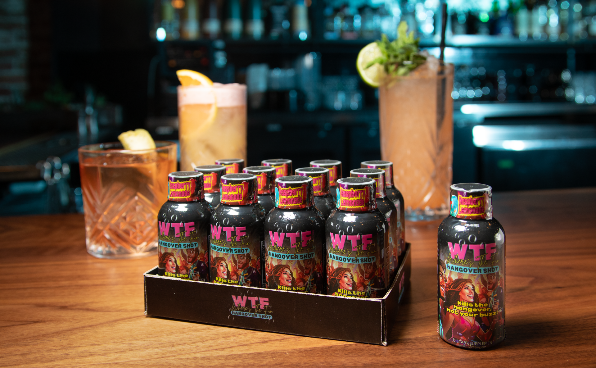 A black box of WTF-labeled cocktail mixer bottles sits on a wooden bar next to several prepared drinks.
