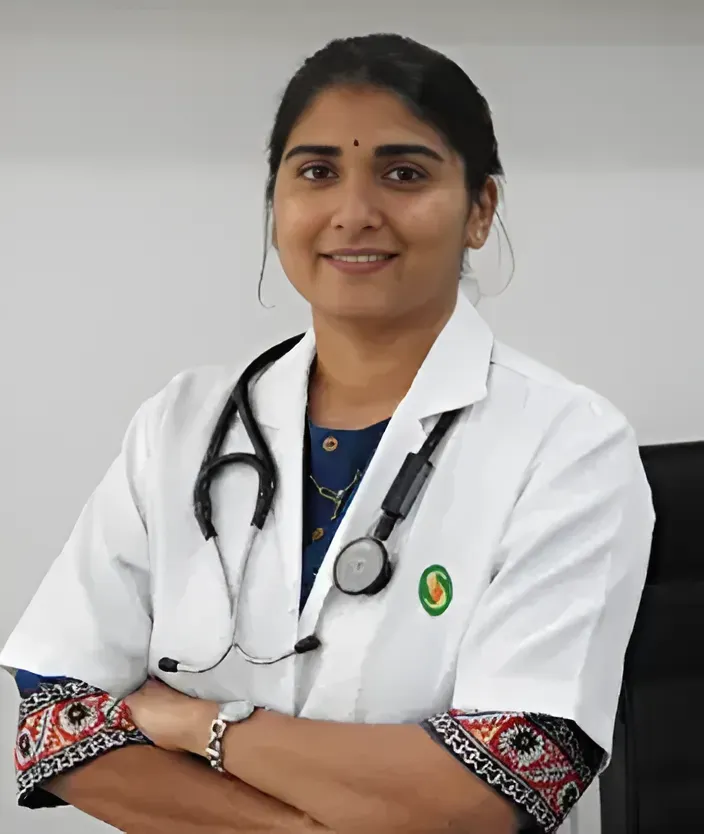 A doctor with a stethoscope around their neck, wearing a white coat, smiling with arms crossed in a professional setting.