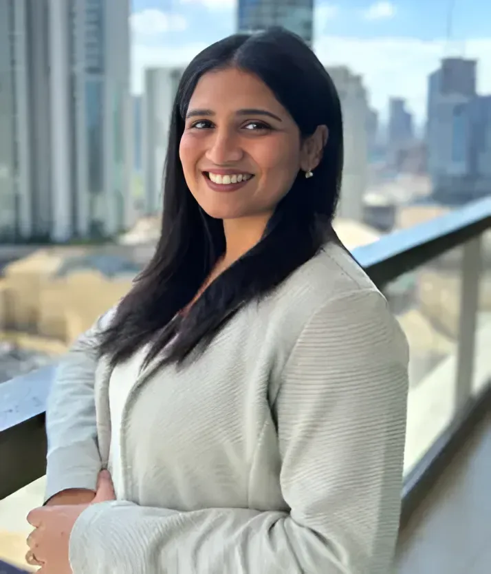 A person smiling at the camera, wearing a light-colored blazer, standing on a balcony with a city skyline in the background.