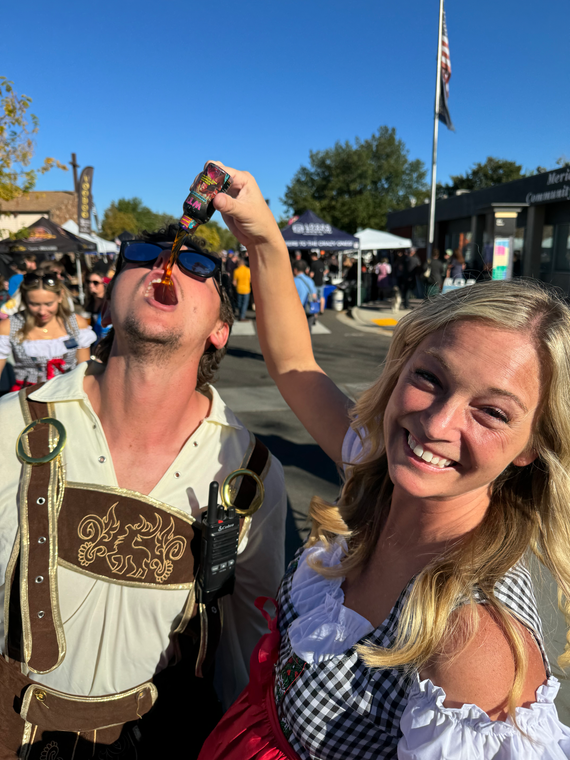 A person in a Bavarian outfit pours a small bottle of liquor into a smiling person's mouth outdoors at a festival.