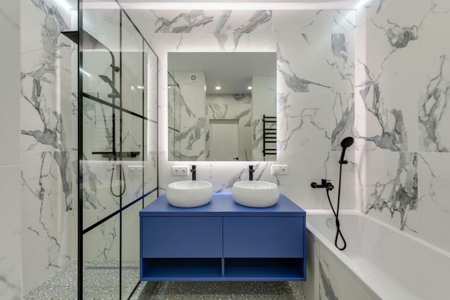 Modern bathroom with marble walls, blue vanity, double sinks, mirror, and glass shower enclosure