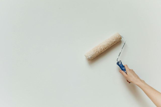 A hand uses a blue-handled paint roller on a white wall, with a section of beige textured paint above.