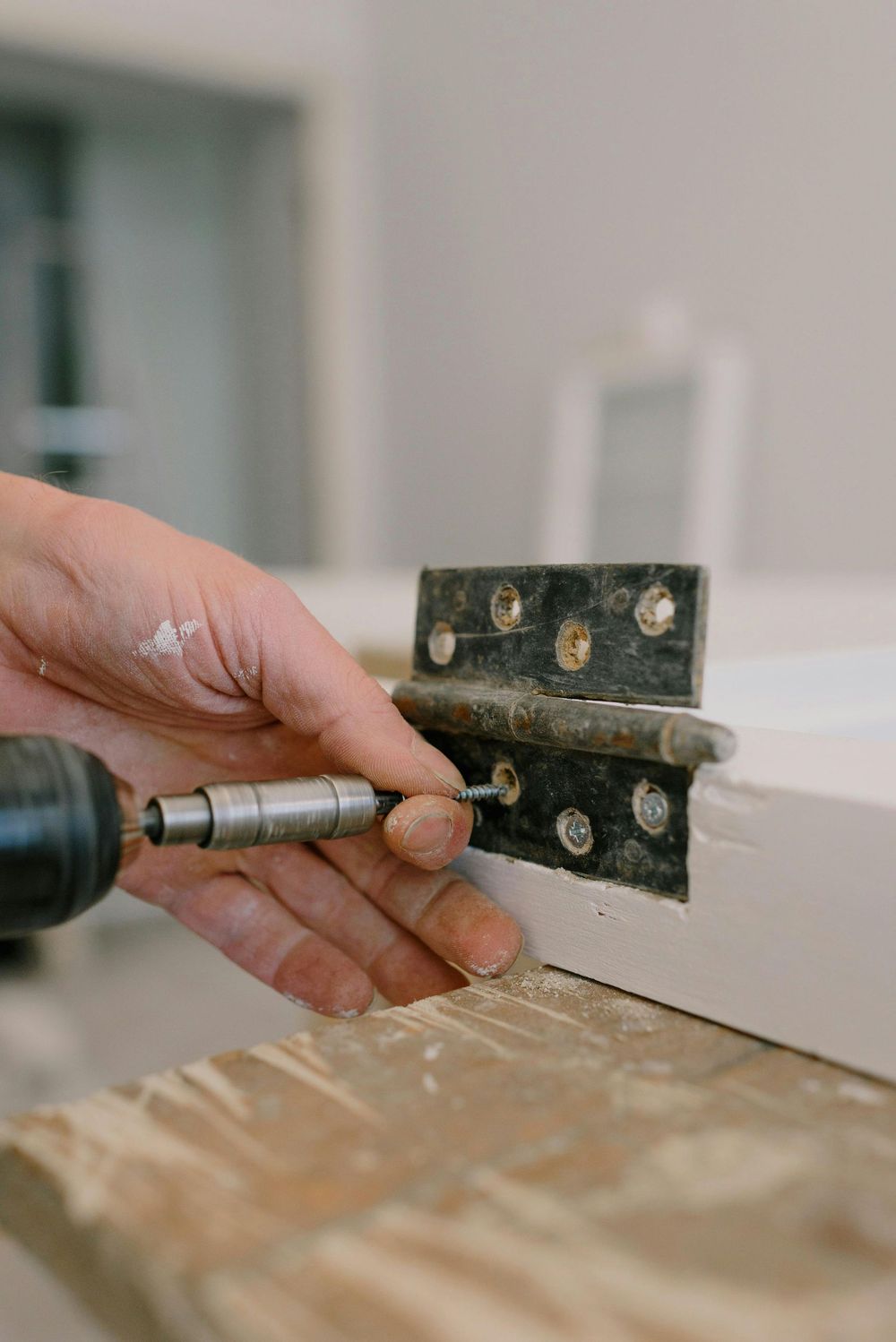 Hand using a drill to drive a hinge into a white cabinet door with wood filler nearby