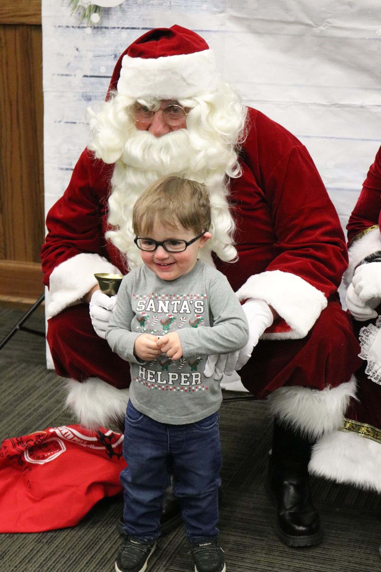 A little boy is sitting next to santa claus holding a bell.