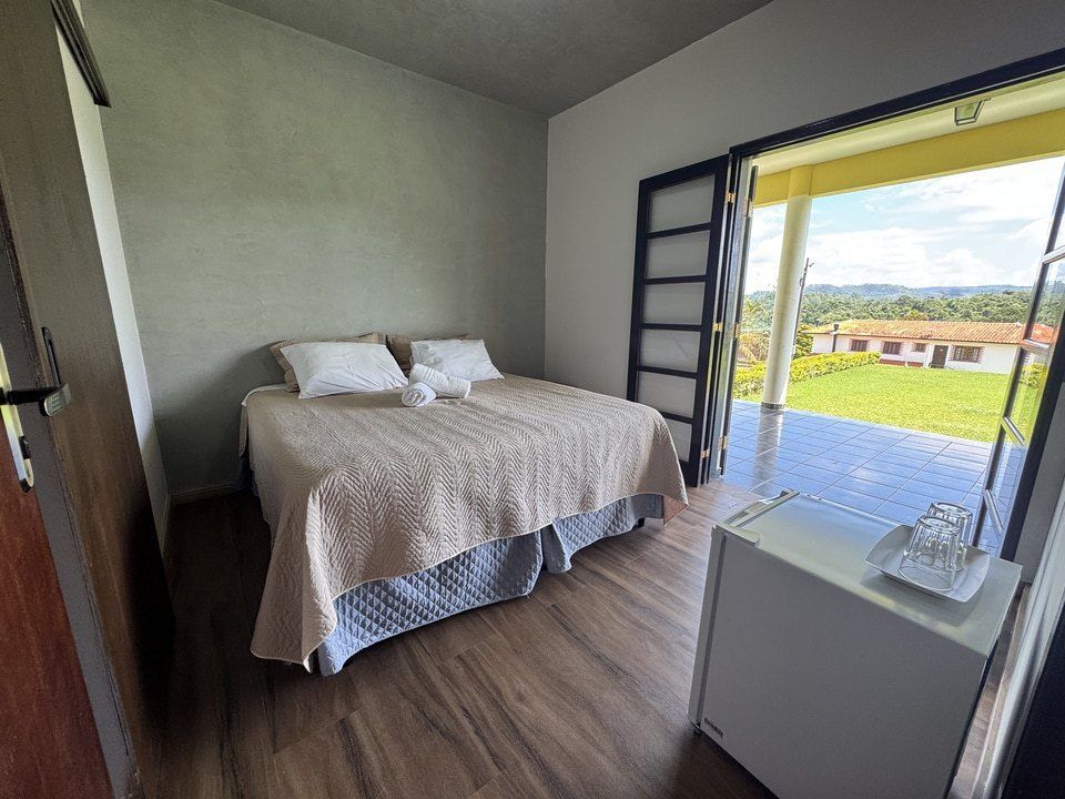 Quarto com cama, porta aberta para um pátio e um pequeno frigobar.