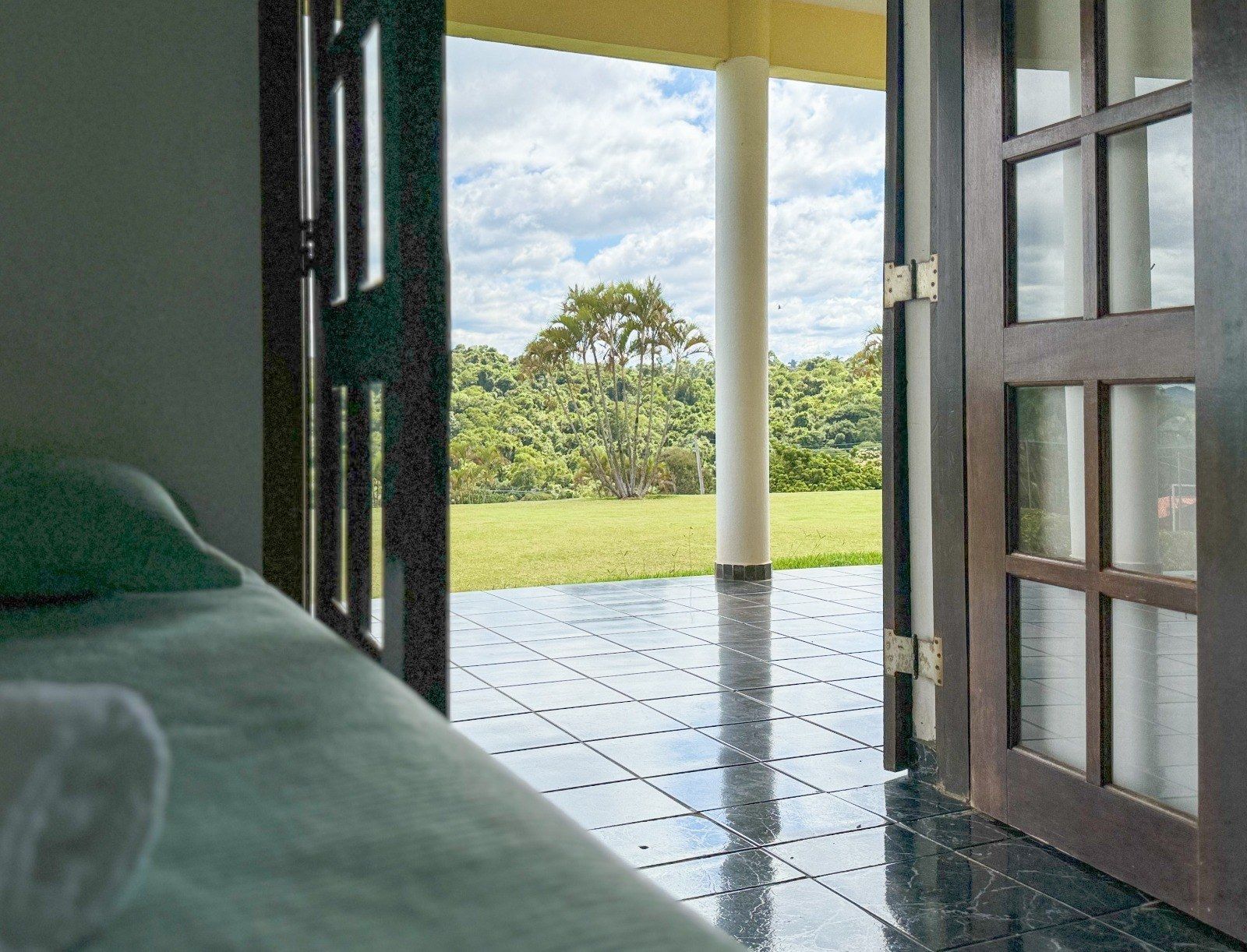 Vista de um campo verde e árvores a partir de um quarto com porta de madeira aberta e uma cama.