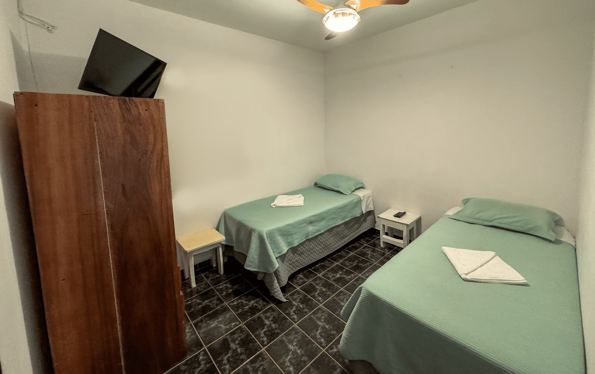 Quarto de hotel pequeno com duas camas de solteiro cobertas com lençóis verdes, piso de azulejos pretos, guarda-roupa de madeira e TV pequena.