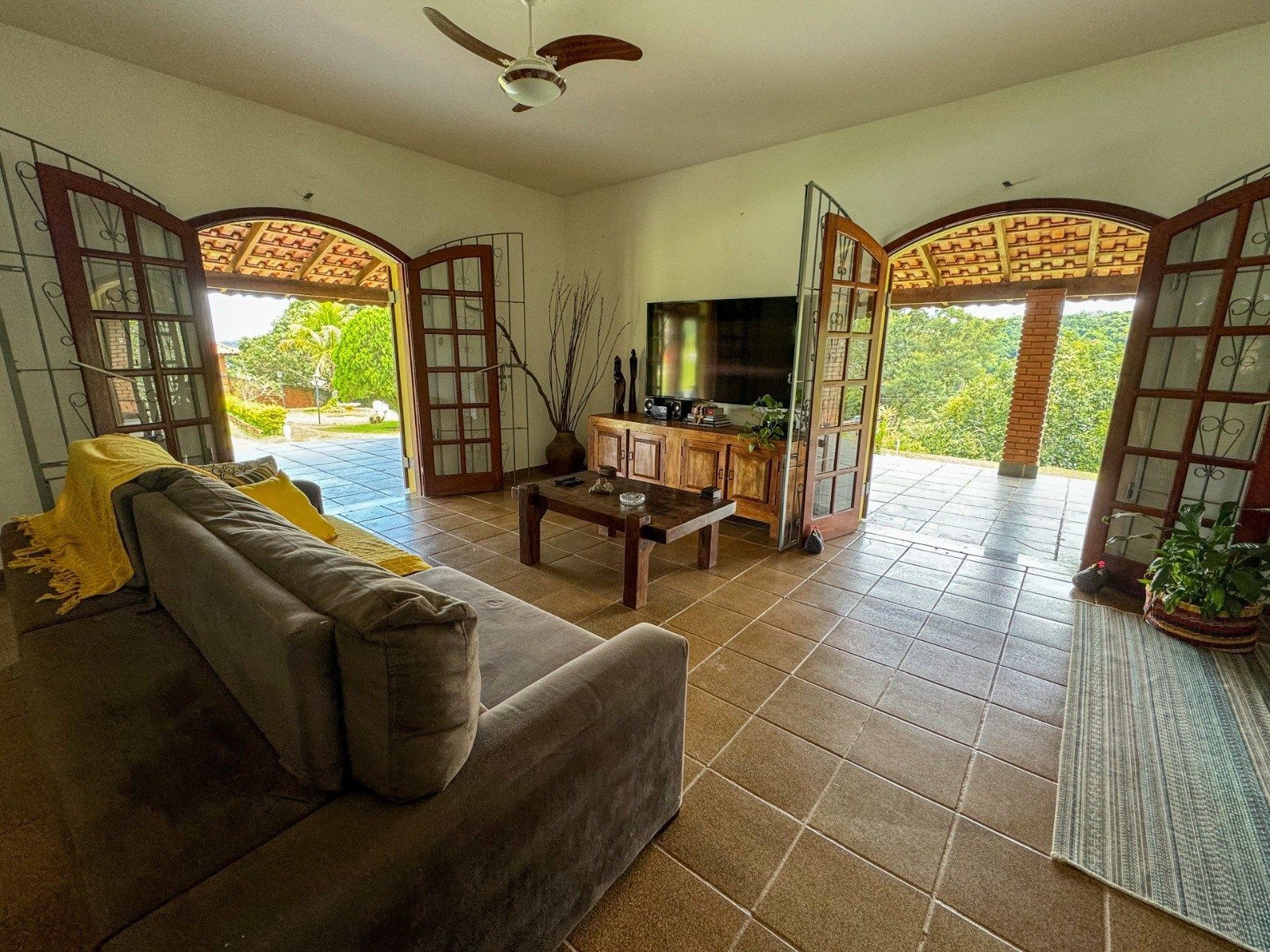 Sala de estar com portas abertas para o exterior, sofá marrom, móveis de madeira e piso de azulejo.