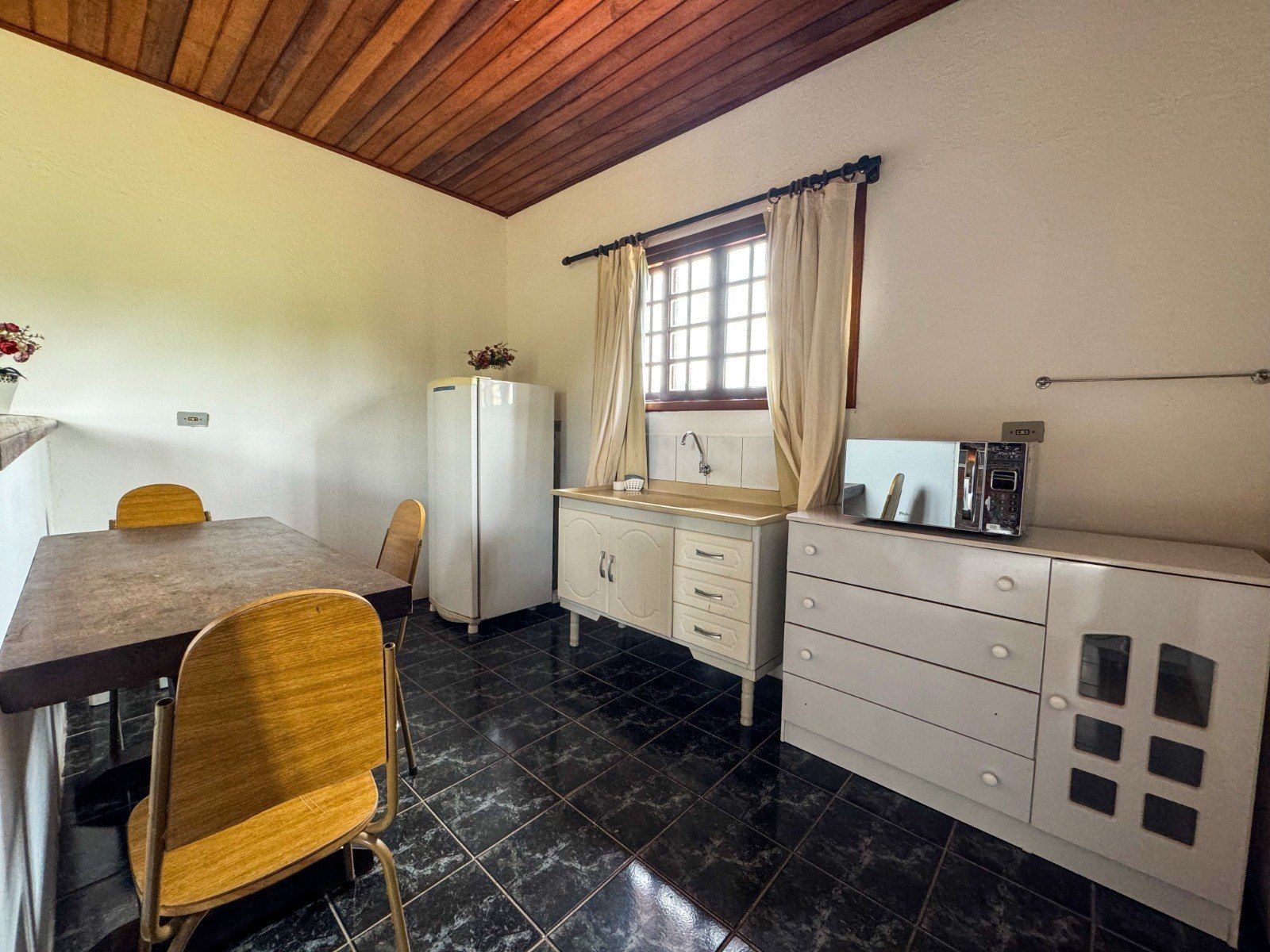 Cozinha com mesa, armários, geladeira e janela. Cadeiras amarelas, piso escuro e teto de madeira.