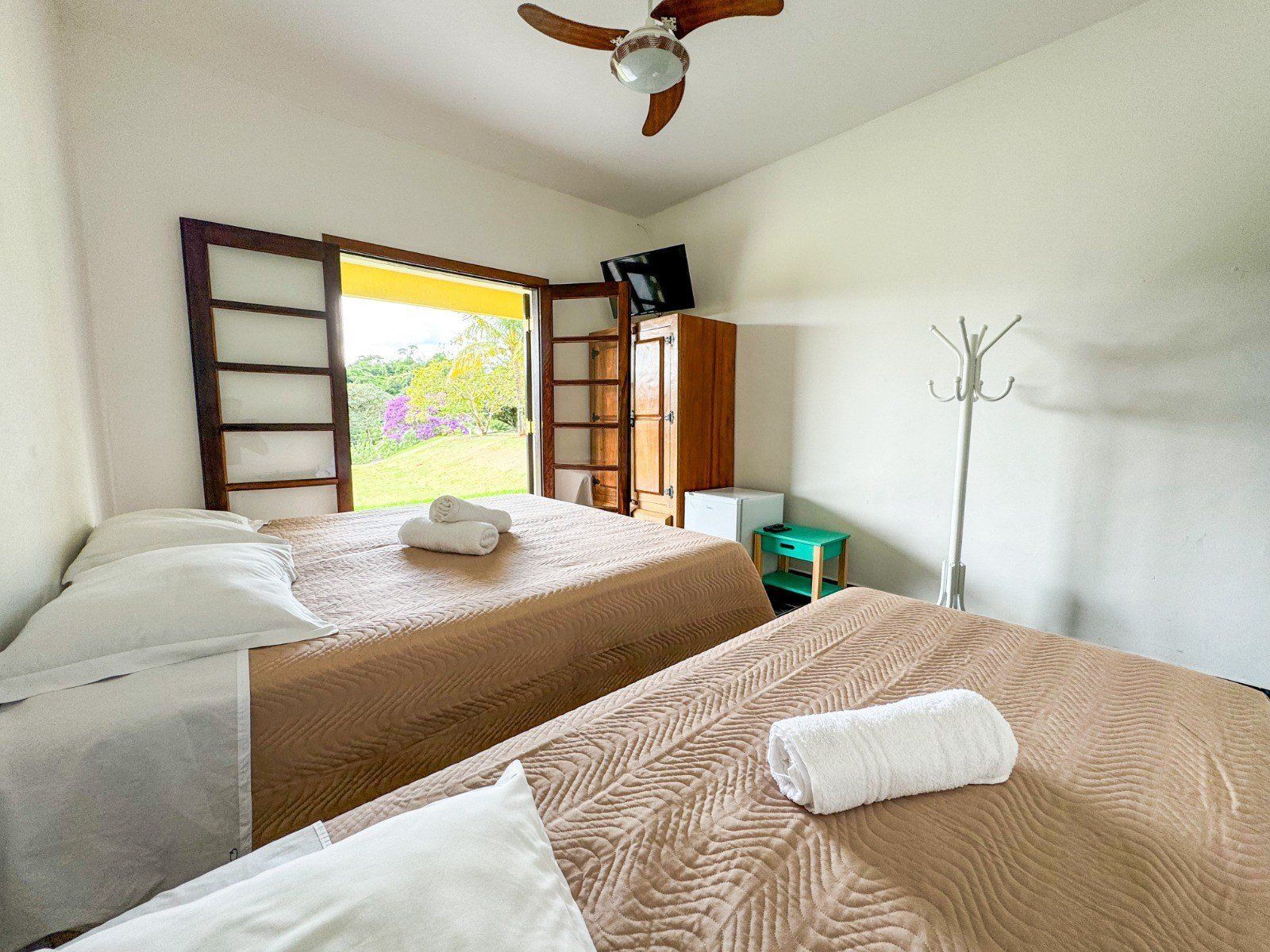 Quarto com duas camas, portas abertas com vista para a vegetação e ventilador de teto.