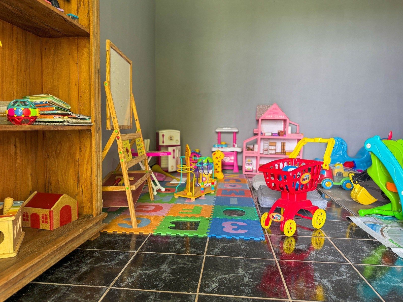 Sala de jogos com brinquedos sobre um tapete colorido; uma prateleira de madeira à esquerda.