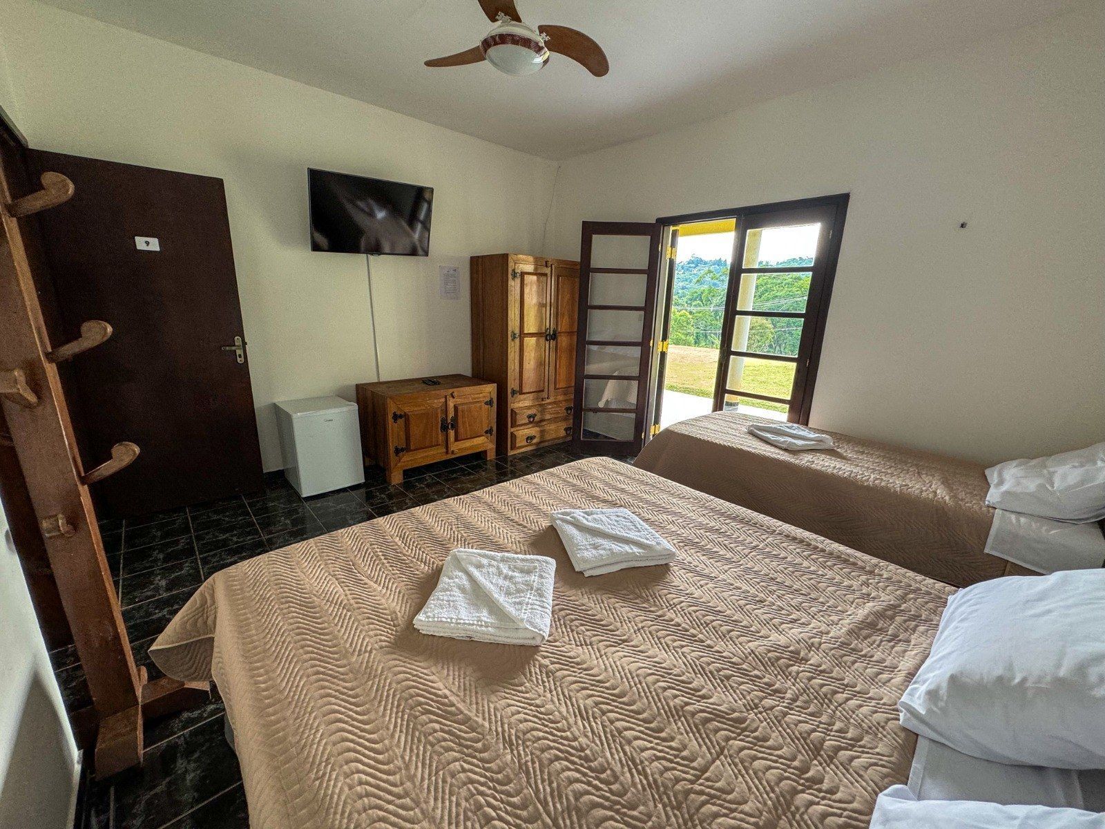 Quarto com uma cama de casal e uma de solteiro, TV, armário, porta com vista, roupa de cama branca.