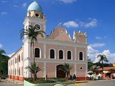 Igreja rosa com cúpula azul e torre do relógio contra um céu azul. Palmeiras e outros edifícios ao fundo.
