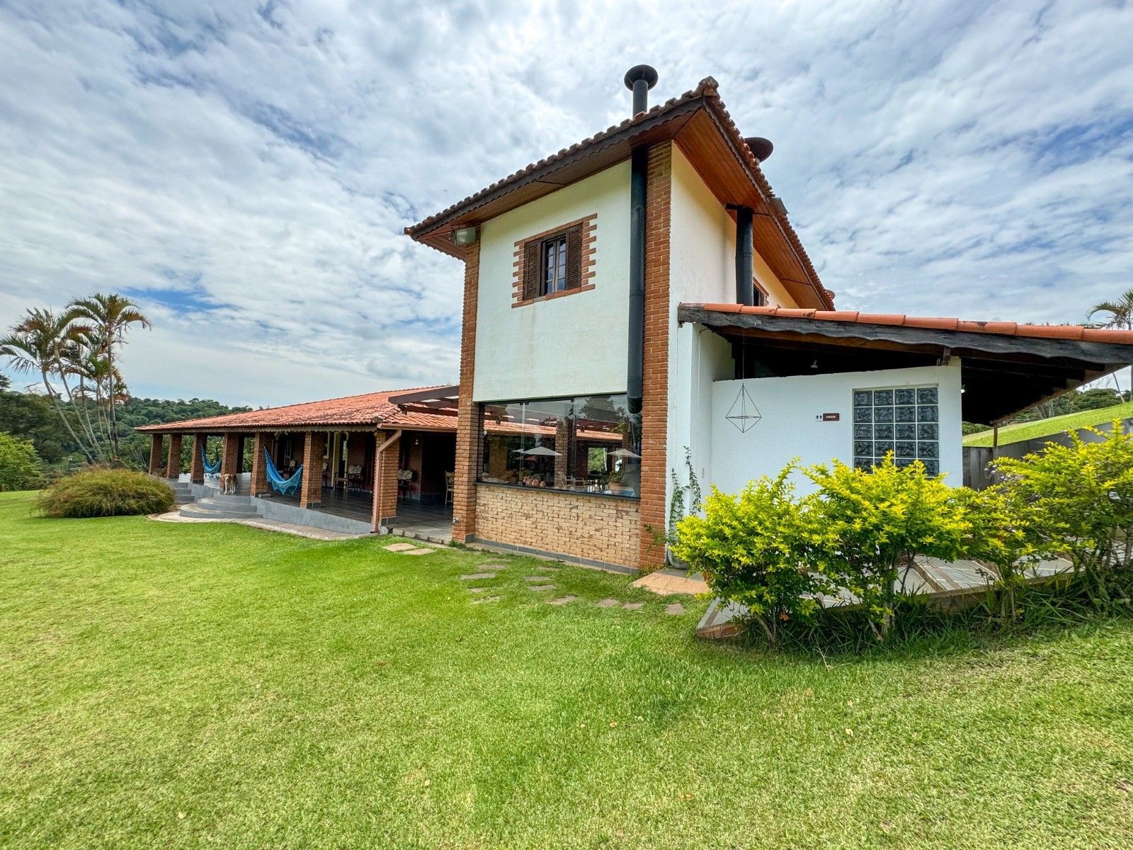 Casa de dois andares com telhado de telha vermelha, paredes brancas e um grande gramado verde sob um céu nublado.