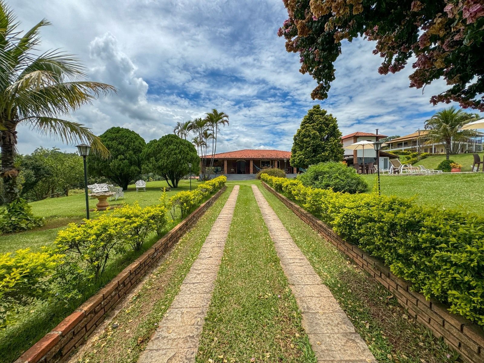 Longa entrada de automóveis que leva a uma casa com telhado de telha vermelha, ladeada por sebes e gramado verde, sob um céu nublado.