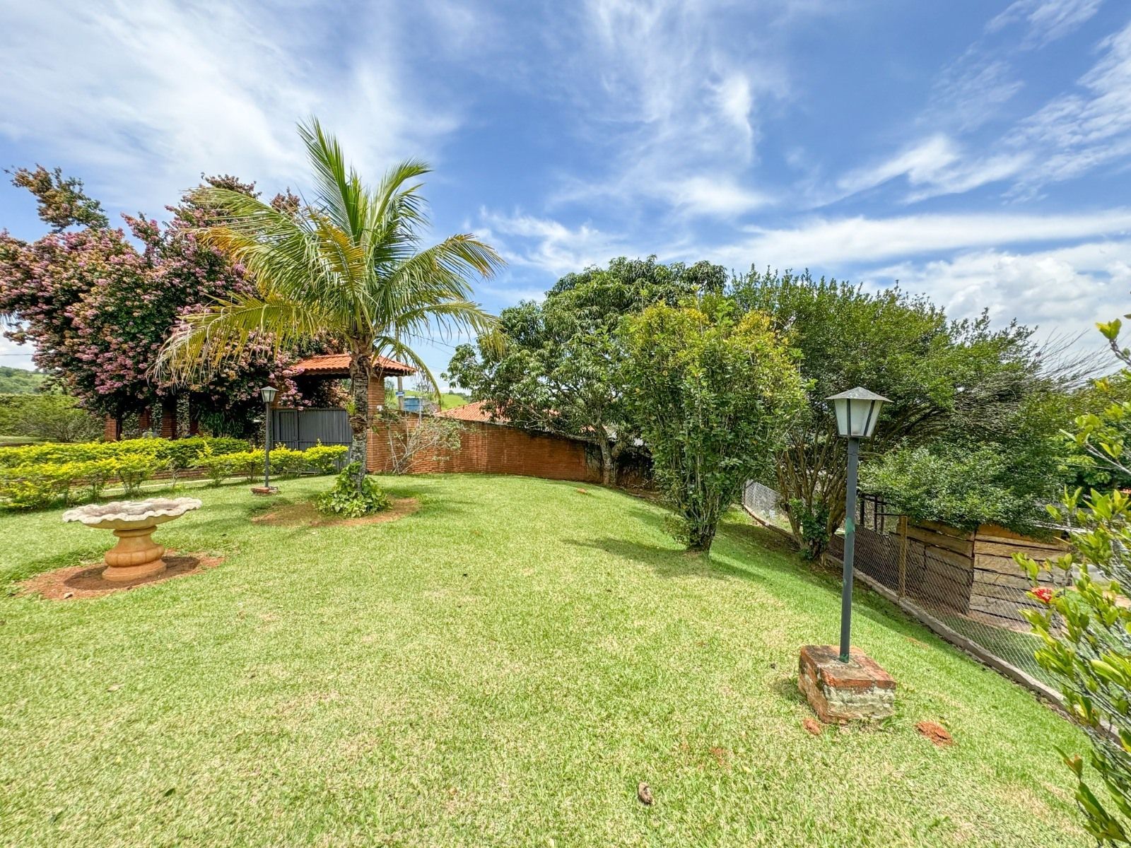 Gramado verdejante com árvores, palmeiras e um poste de luz, sob um céu azul com nuvens.