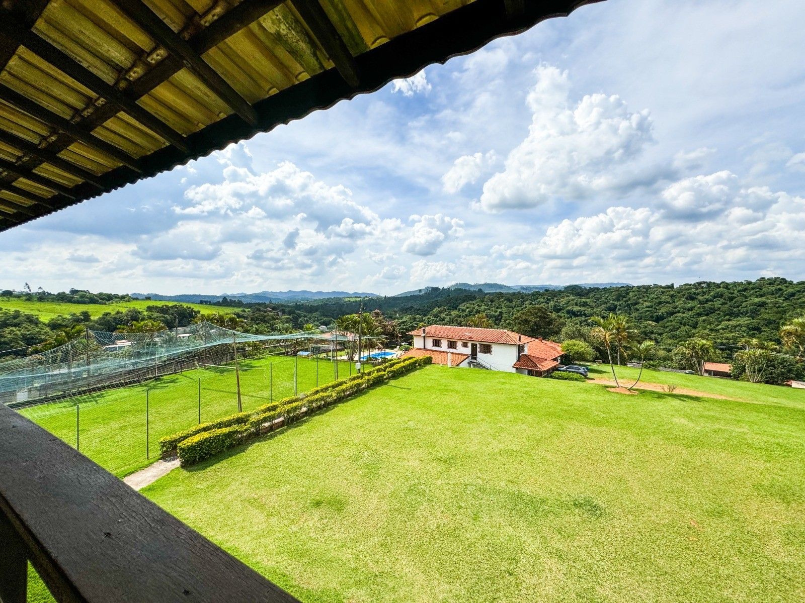 Paisagem verdejante com uma casa e quadras de tênis sob um céu nublado, vista de uma varanda de madeira.