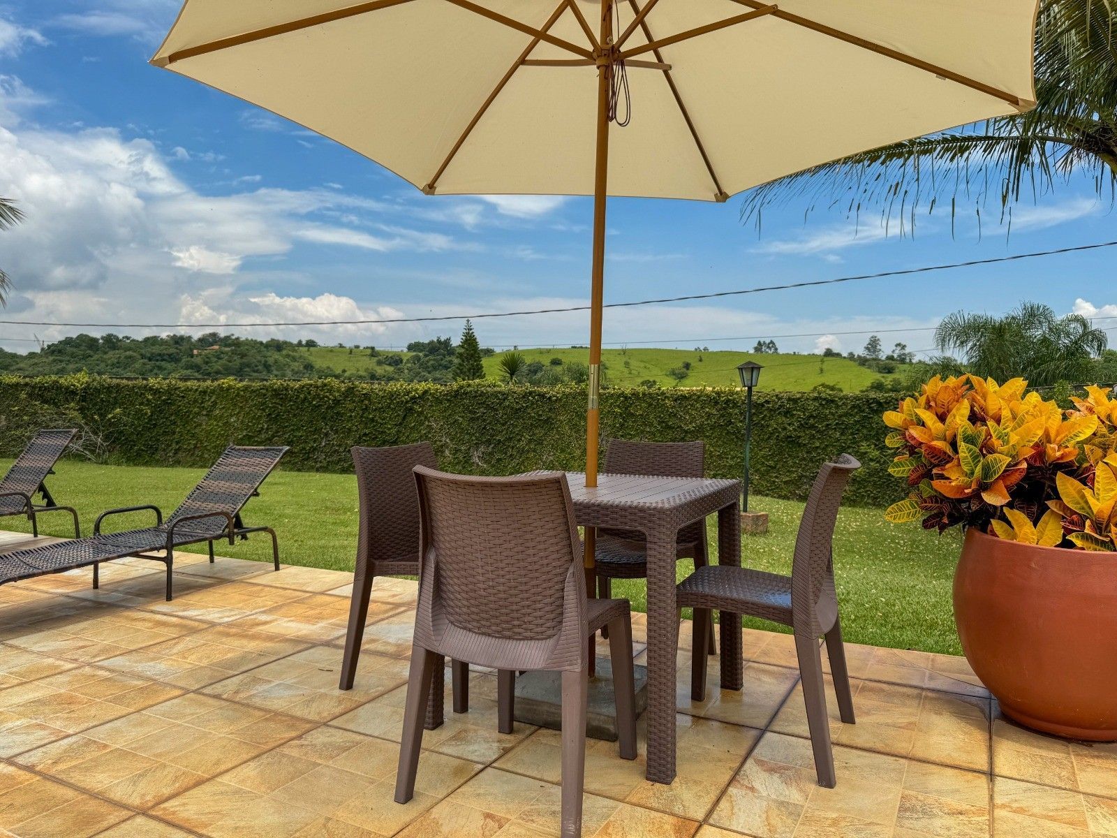 Pátio com mesa, cadeiras, guarda-sol e vasos de flores, com vista para um gramado verde sob um céu azul.