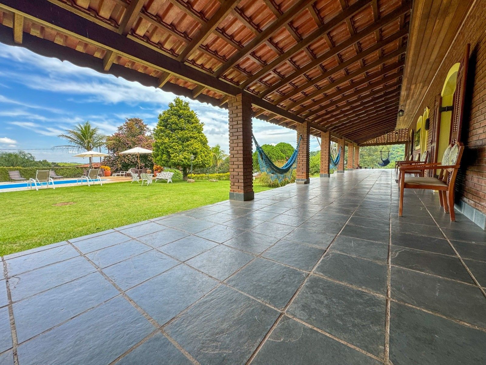 Varanda coberta com piso de azulejo, com vista para um gramado, árvores e uma piscina. Rede pendurada. Céu azul.