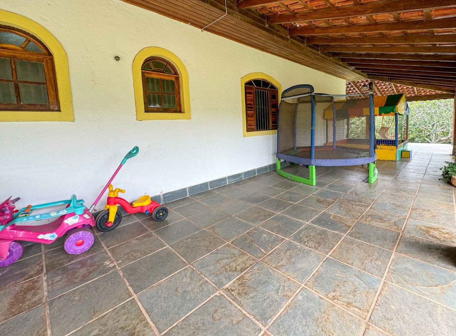 Varanda coberta com brinquedos infantis, incluindo um carrinho rosa, um triciclo e uma cama elástica.