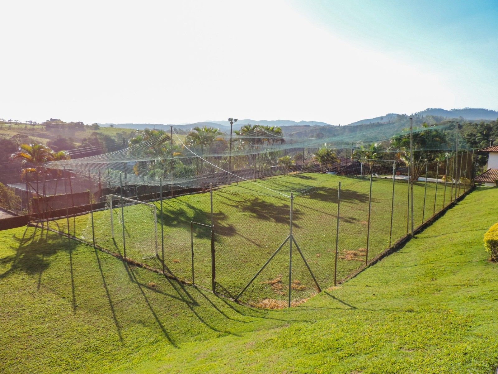 Encosta verdejante com uma cerca de arame em volta de uma estrutura de rede; árvores e colinas ao fundo.