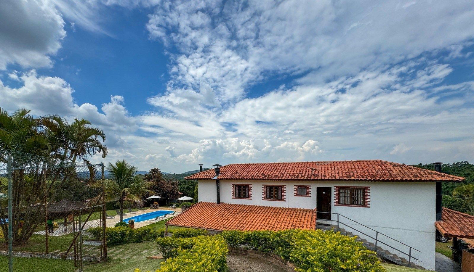 Casa branca com telhado laranja, piscina e palmeiras sob um céu azul parcialmente nublado.