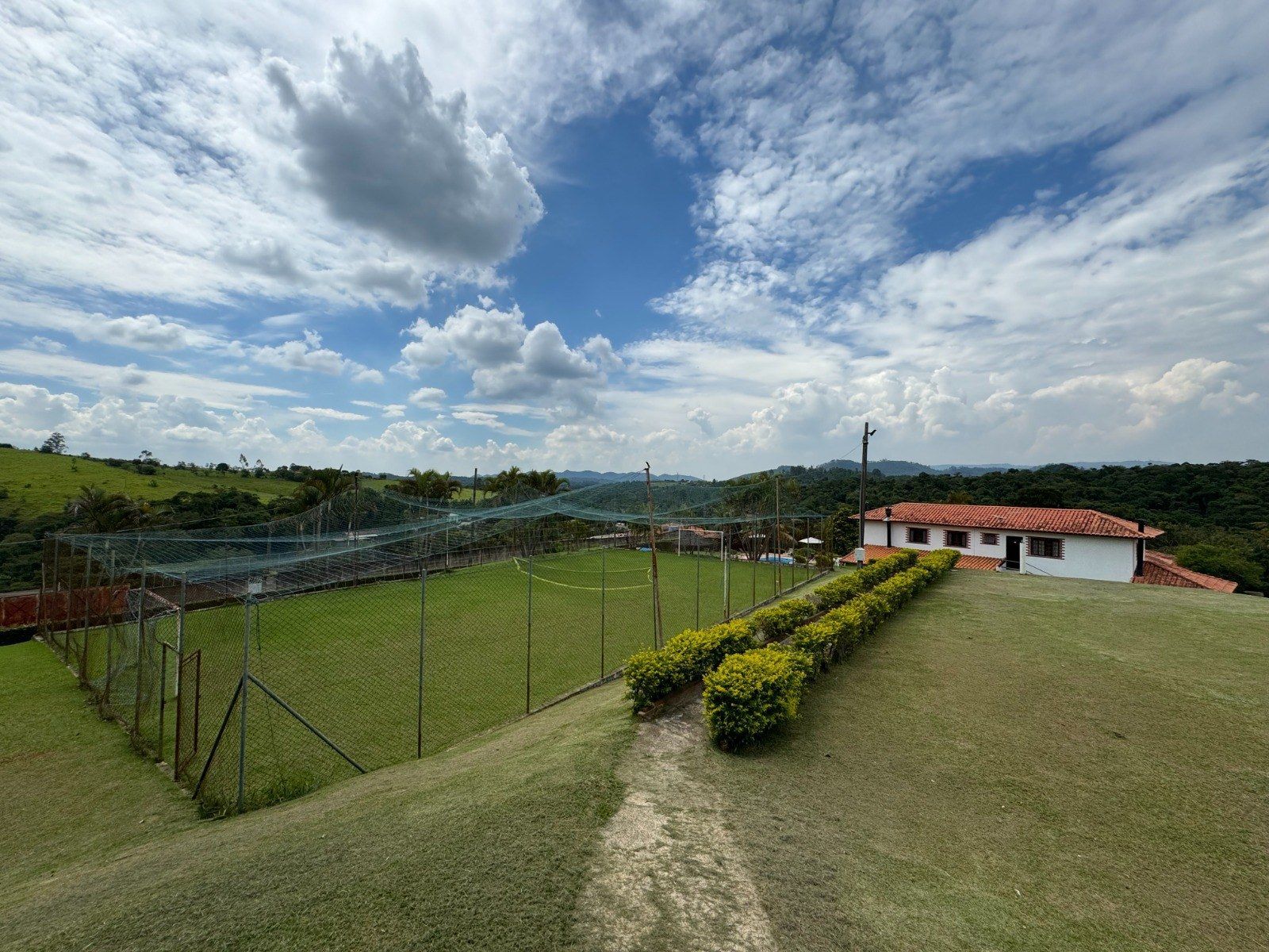 Campo verde com um muro baixo, uma longa sebe, um edifício branco e céu azul com nuvens fofas.