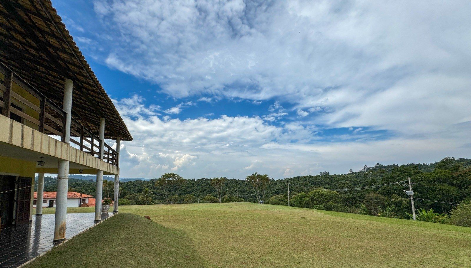 Prédio de dois andares com varanda com vista para um campo gramado e árvores verdejantes sob um céu nublado.