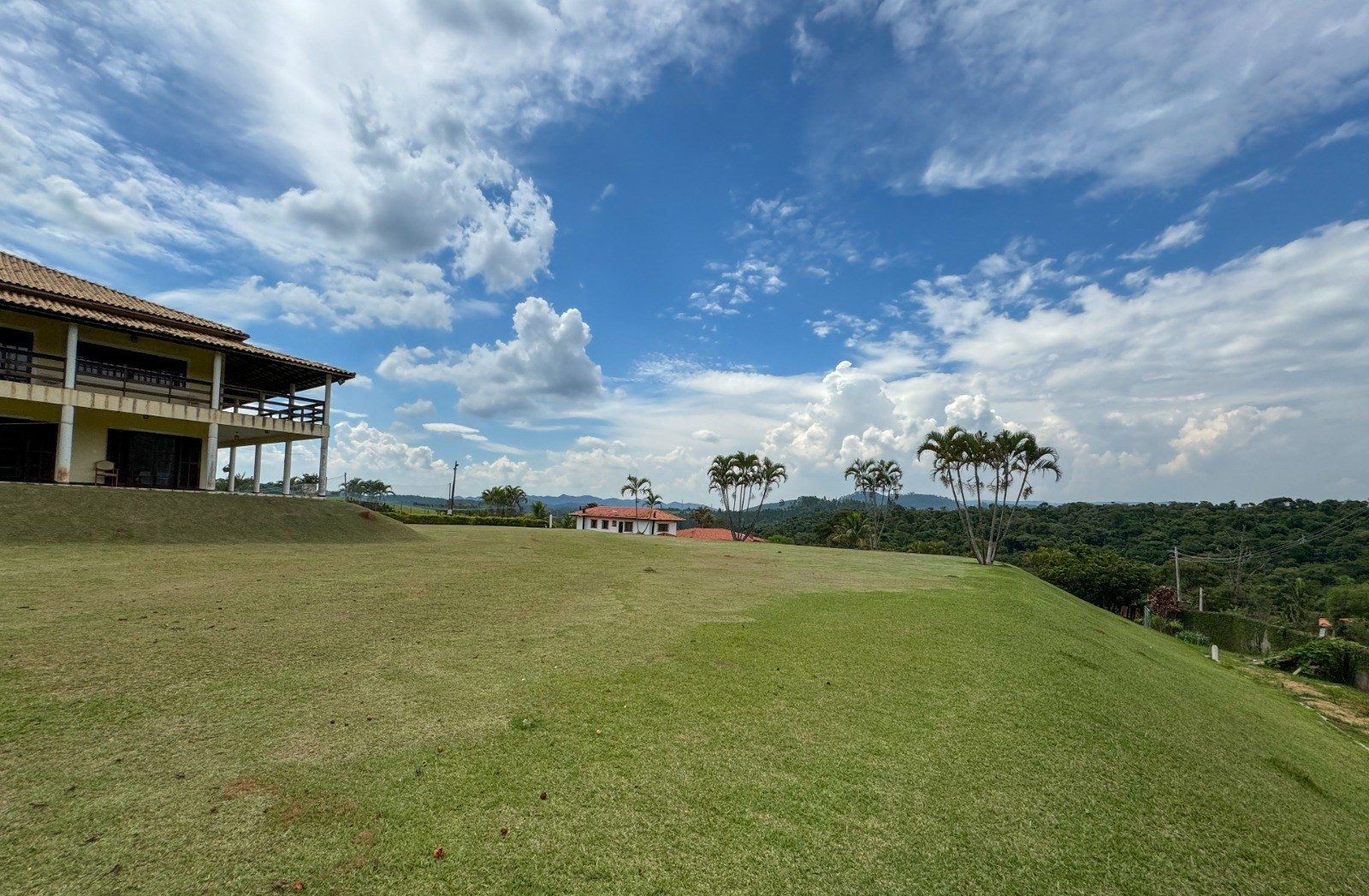 Quintal verdejante com uma casa de dois andares, uma casa menor e árvores sob um céu azul com nuvens.