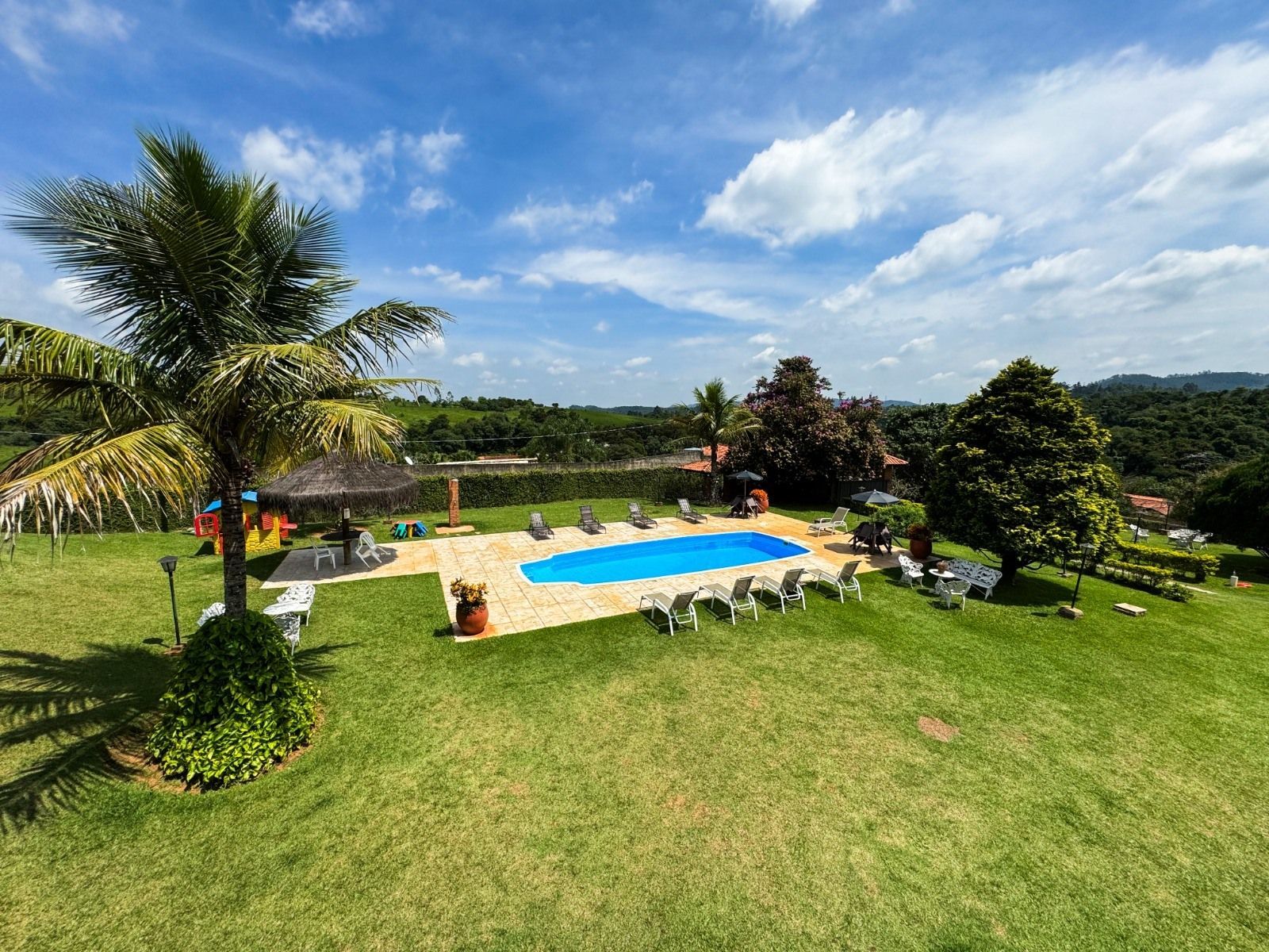 Piscina rodeada por relva verde, árvores e espreguiçadeiras sob um céu azul com nuvens brancas.