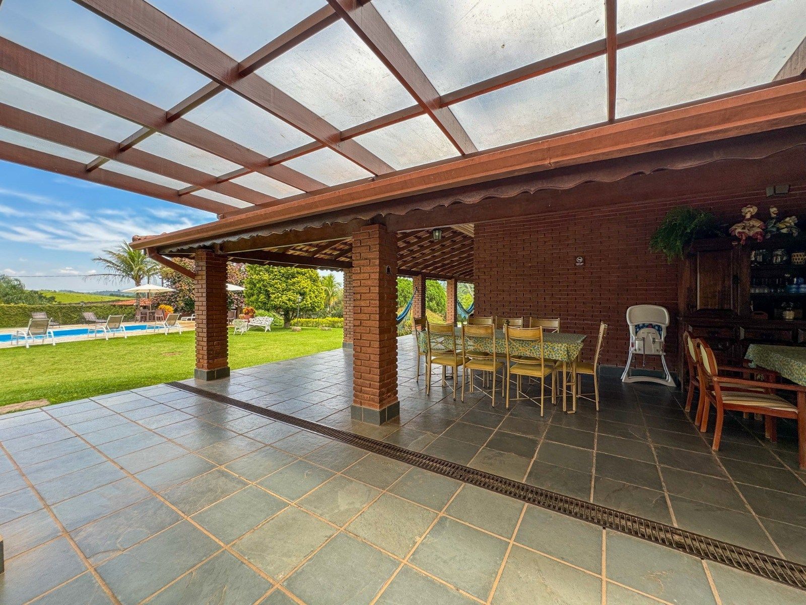 Pátio coberto com mesa de jantar, colunas de tijolos e vista para a piscina e o jardim.