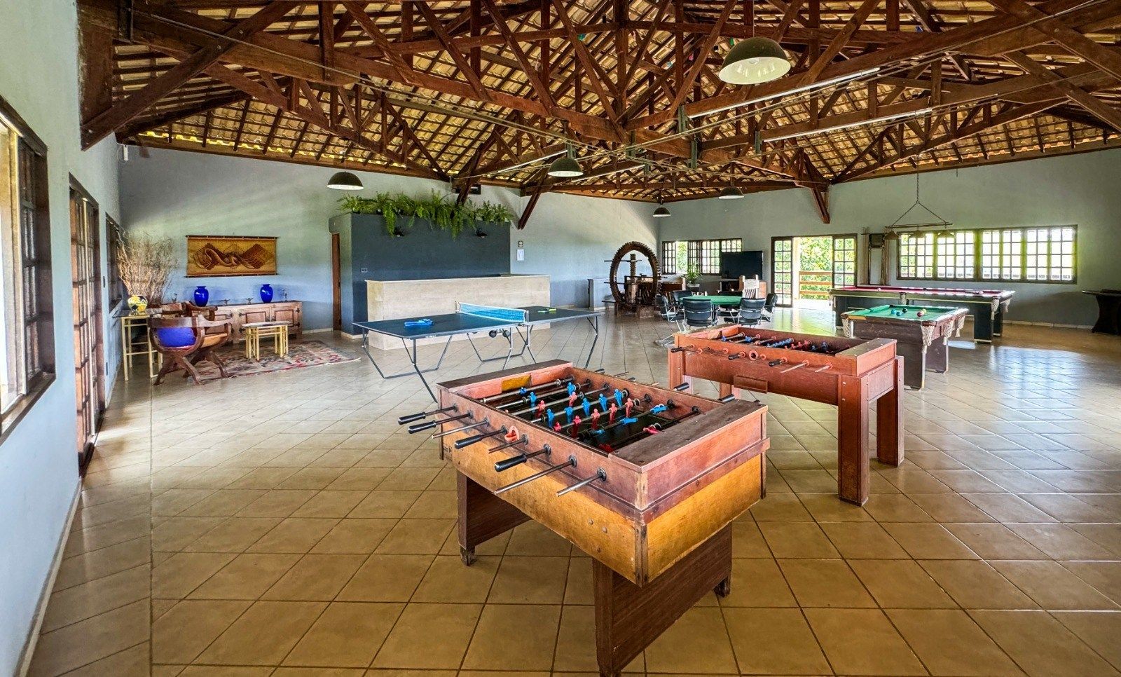 Mesas de pebolim em uma ampla sala de recreação com bilhar, mesa de jantar e área de bar, com teto alto de madeira.