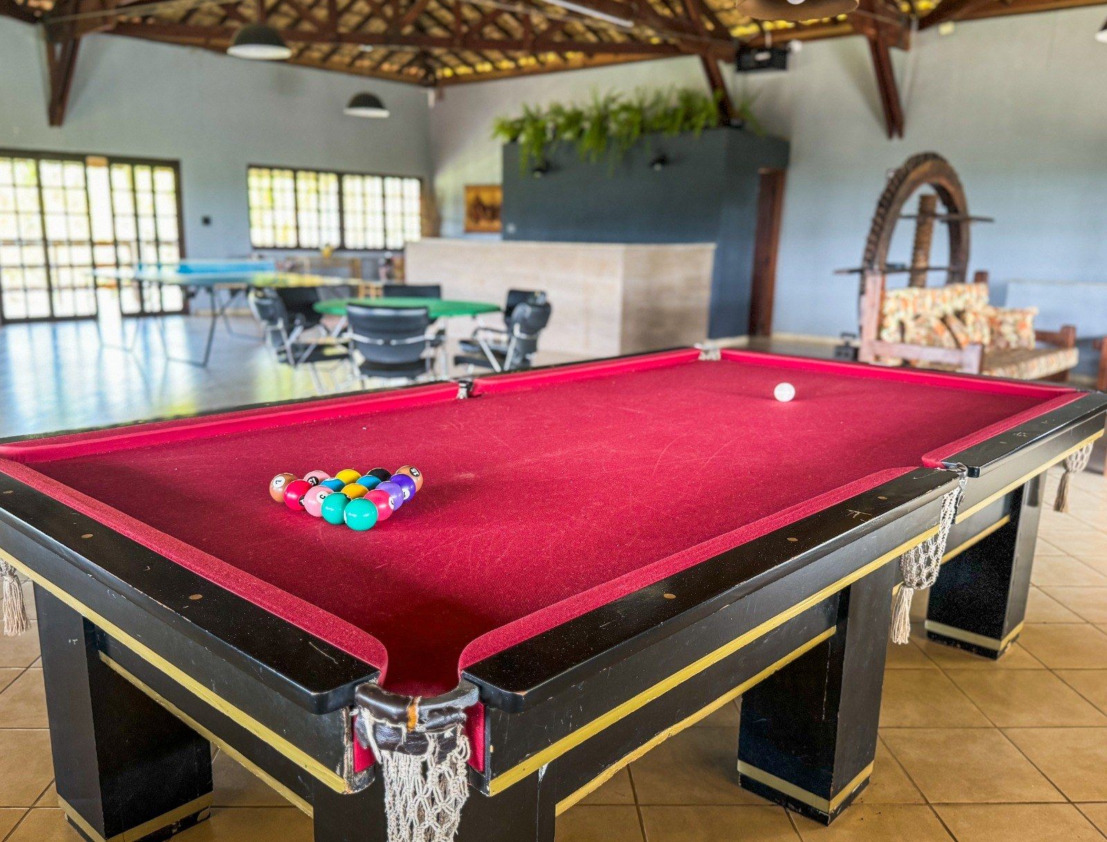 Mesa de bilhar com bolas enfileiradas, feltro vermelho, em uma área de lazer. Outras mesas de jogos são visíveis.