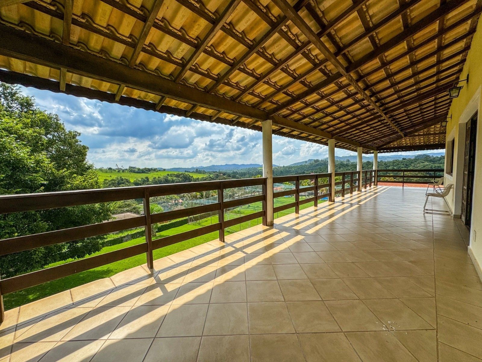 Varanda coberta com vista para uma paisagem verdejante, com corrimão de madeira e piso de azulejo.