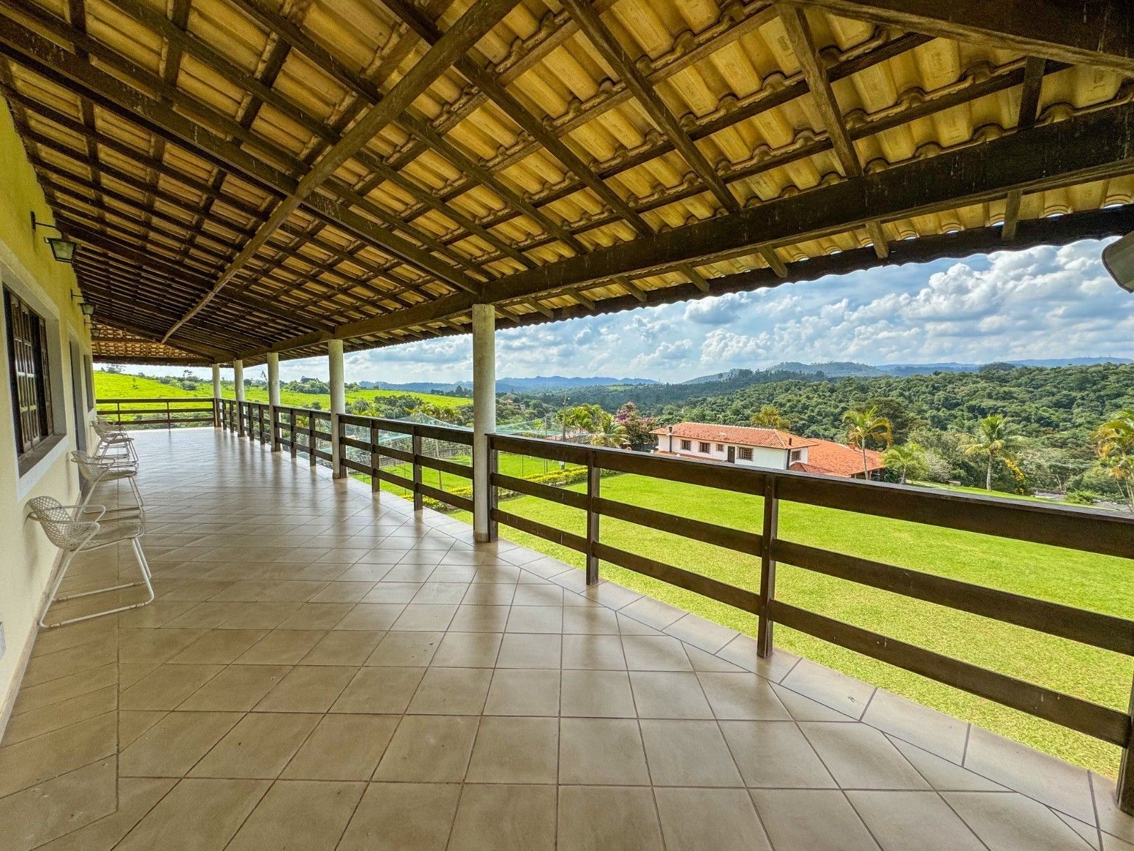 Varanda espaçosa com piso de azulejo, corrimão de madeira e vista para a paisagem verdejante e as árvores.