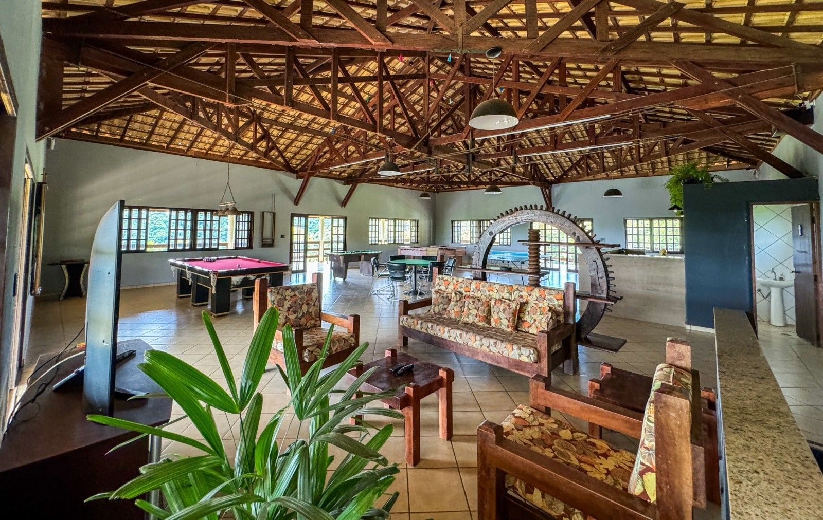Interior espaçoso com vigas de madeira expostas, sofás, mesa de bilhar e uma grande roda mecânica.