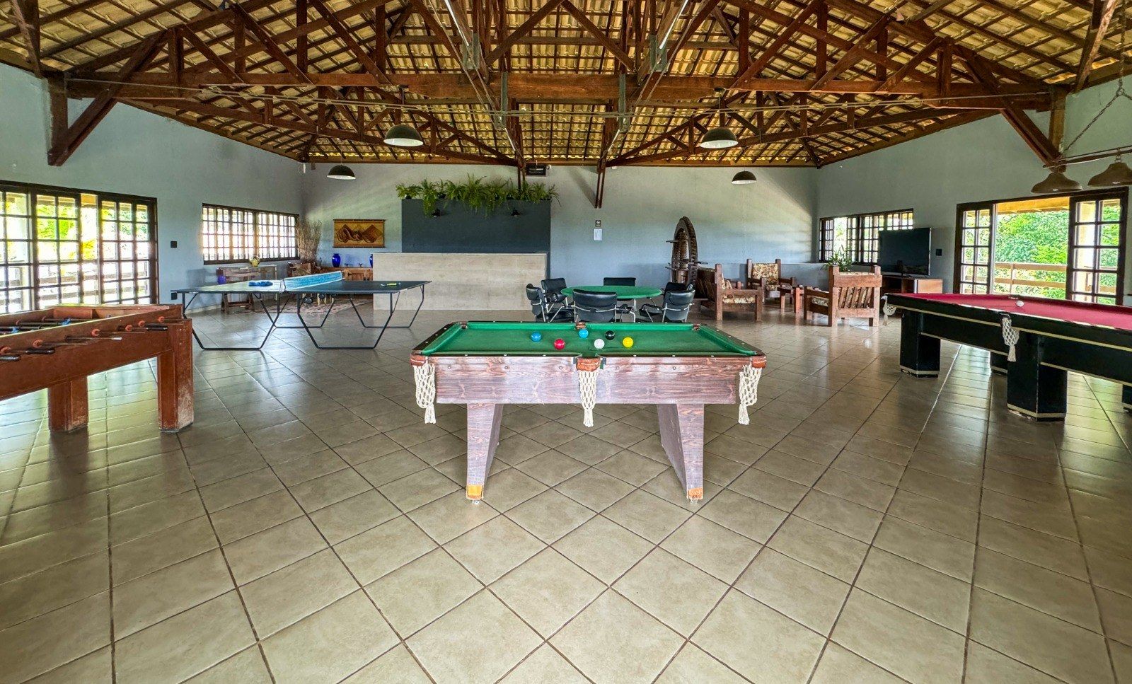 Sala de jogos com mesa de bilhar, pebolim e pingue-pongue, com janelas e teto com vigas de madeira.