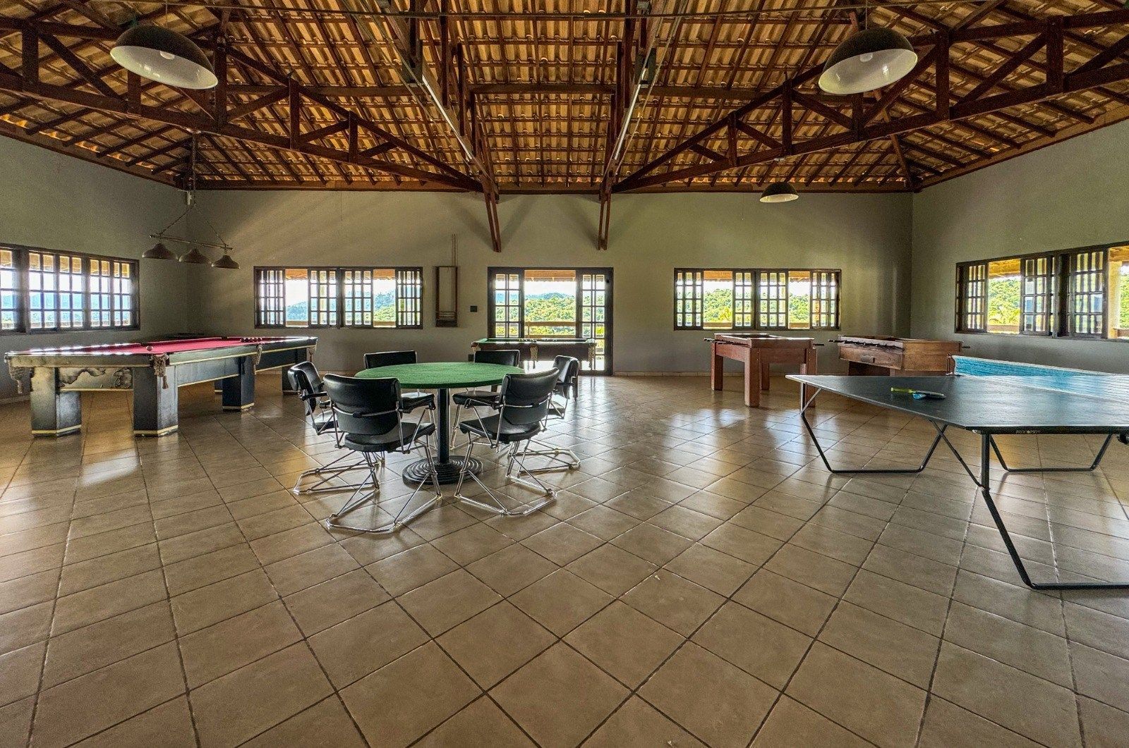 Sala de jogos com mesa de bilhar, mesa de pingue-pongue e mesas de jantar, janelas e piso de azulejo.