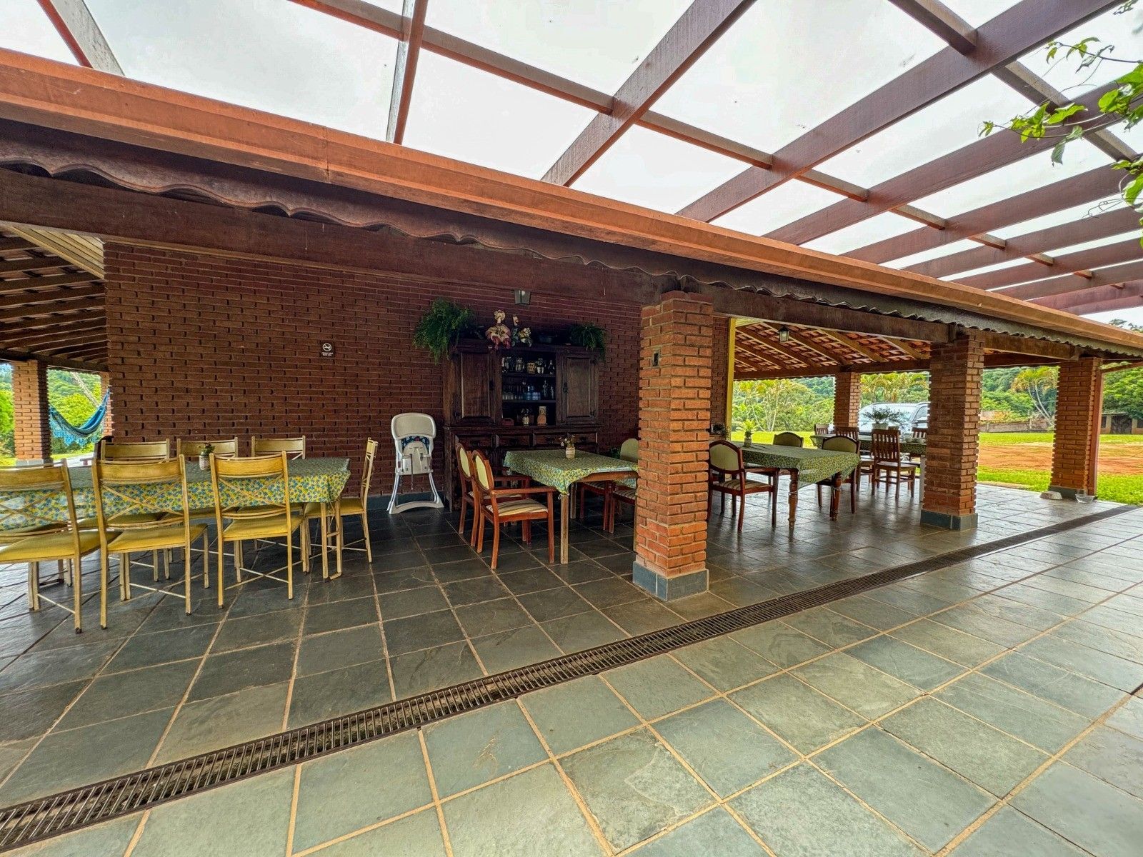 Área de jantar externa com paredes de tijolos, mesas e cadeiras sob um pergolado de madeira.