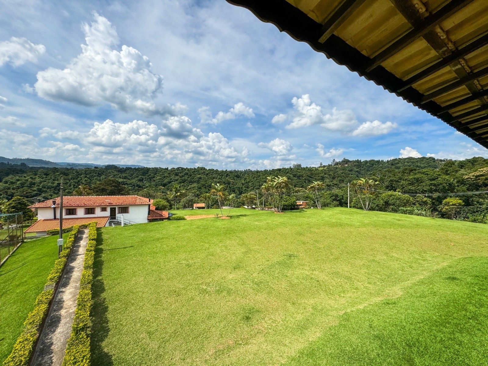 Uma casa branca com telhado de telhas vermelhas fica em uma colina gramada com vista para uma floresta sob um céu azul nublado.
