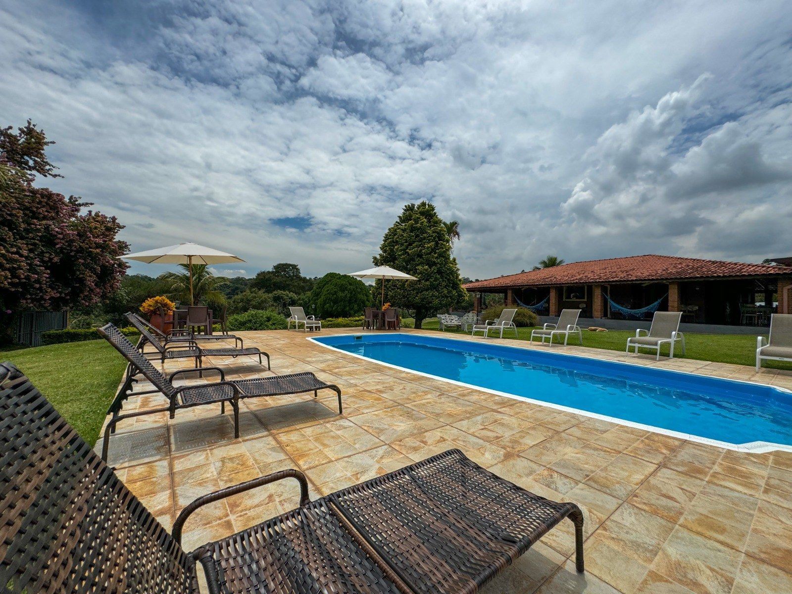 Vista da piscina com espreguiçadeiras, guarda-sóis e prédio sob um céu nublado.
